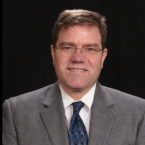 A man in a suit and tie smiles for the camera, conveying an air of professionalism and confidence.