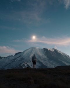 A man standing on top of a mountain making eye contact with the moon.