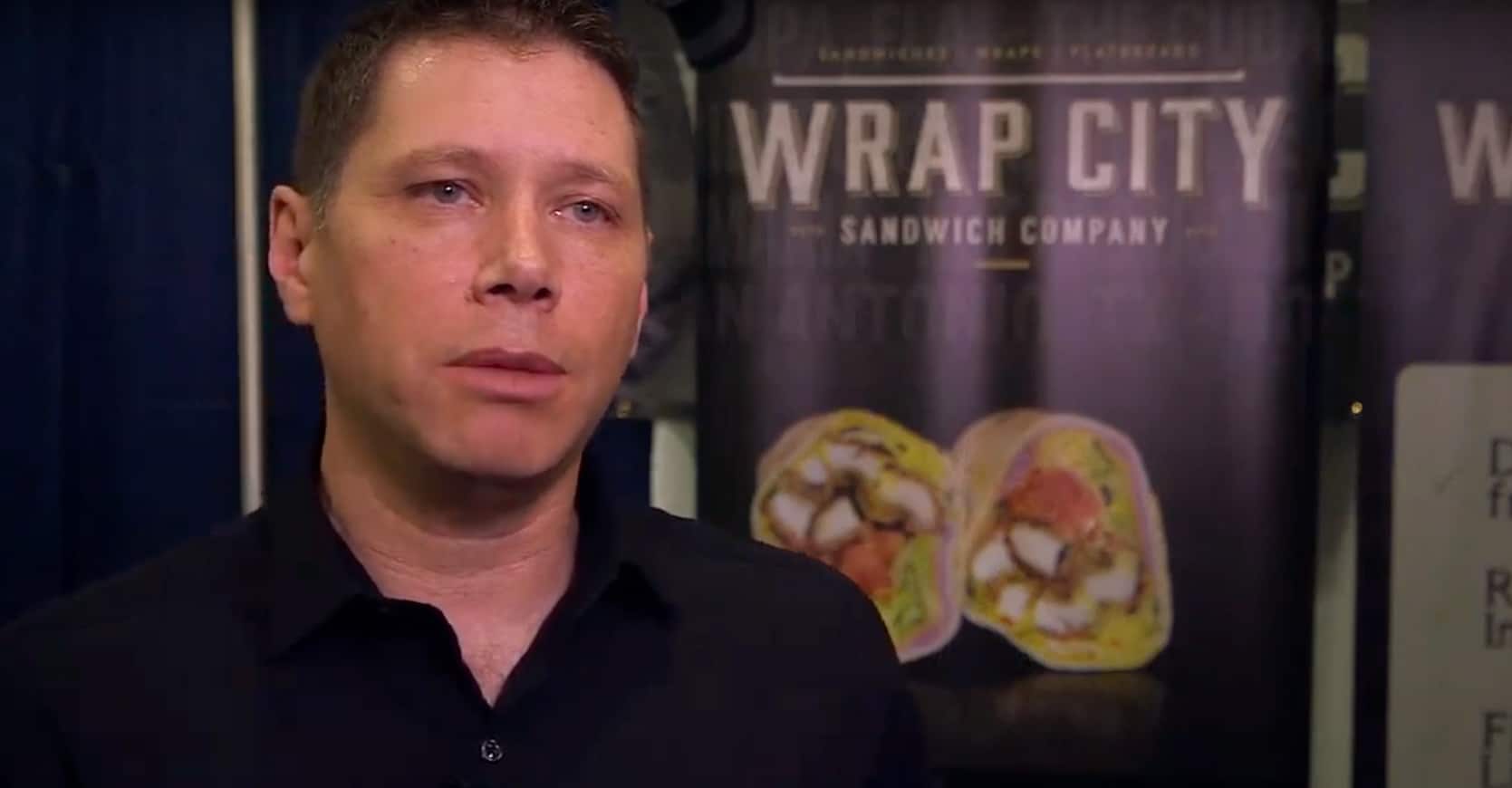 About Description: A man in a black shirt standing in front of a wrap city banner.