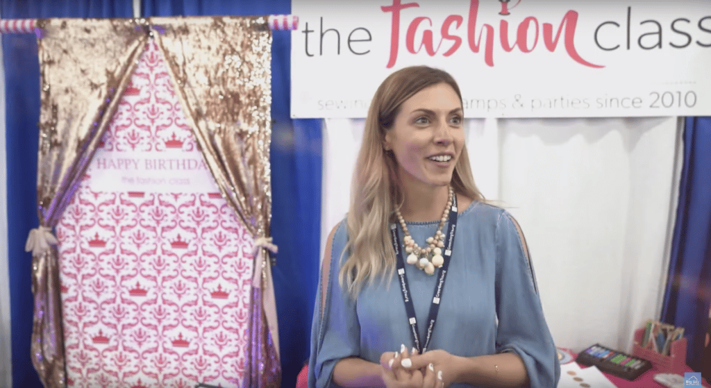 A woman standing in front of a booth at a fashion show, receiving testimonials from impressed attendees.