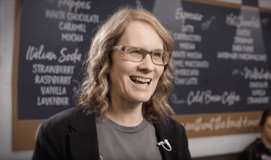 A woman in glasses is smiling while giving testimonials in front of a chalkboard.